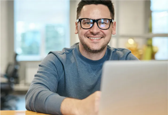 hombre con lentes sonriendo detrás de la pantalla de una computadora portátil